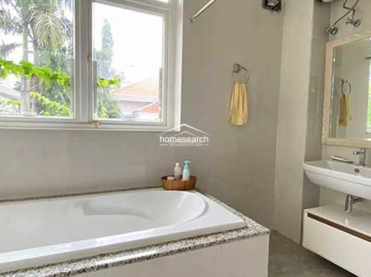 This bathroom features a clean, minimalist design with neutral-toned walls and a large window that invites natural light while offering a view of lush greenery. The space includes a deep soaking bathtub with a granite-style surround and a modern floating vanity with an integrated sink, reflecting a well-maintained and contemporary standard.