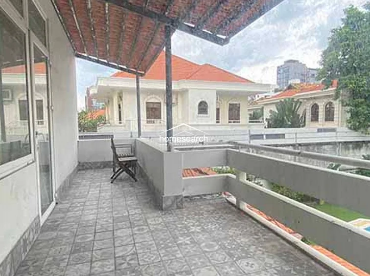 This covered balcony offers a semi-outdoor living space with decorative patterned floor tiles and a rustic, exposed-beam ceiling structure. It provides a peaceful view of the surrounding neighborhood and neighboring villas, serving as a versatile area for relaxation or outdoor dining. The space is well-maintained and connects directly to the interior, extending the home's usable living area into the open air.