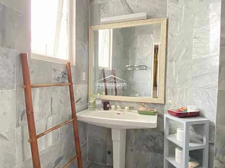 This bathroom features floor-to-ceiling grey marble-effect tiling and a pedestal sink with a large, ornate-framed mirror. A bamboo ladder serves as a rustic towel rack, while a small grey storage unit provides additional space for toiletries.
