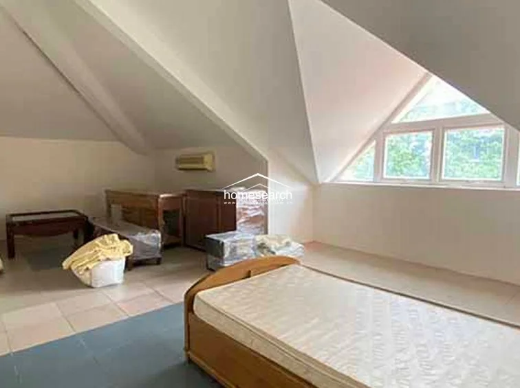 This attic-style bedroom features a dramatic vaulted ceiling and a large triangular window that floods the space with natural light. The room is sparsely furnished with a wooden bed frame and a small side table, with a wall-mounted AC unit ensuring climate control.