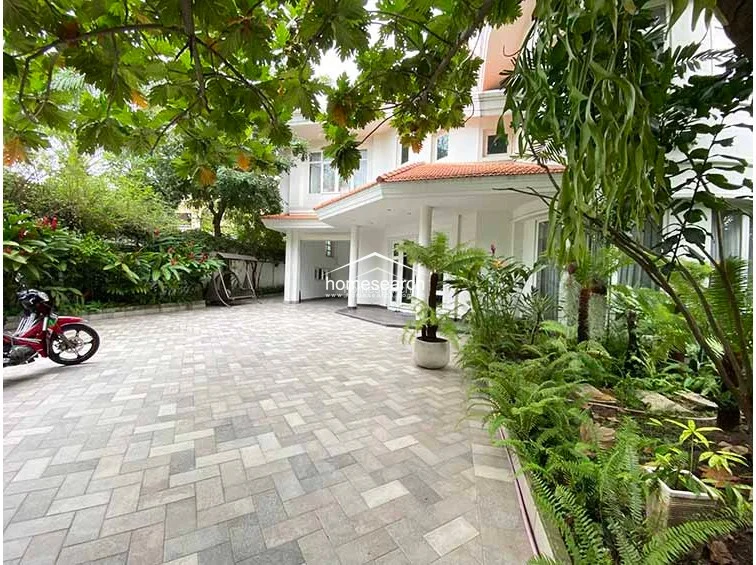 The exterior grounds feature a spacious, paved driveway area finished in a neutral herringbone-patterned stone, providing ample parking space. The area is heavily landscaped with tropical foliage, ferns, and mature trees, creating a private, garden-like entrance that connects directly to the home's main living level.