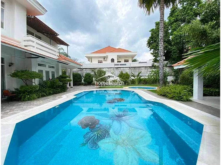 This outdoor space features a large, custom-tiled swimming pool with a decorative floral mosaic at its base, surrounded by a paved stone patio and lush landscaping. The property includes a covered terrace area, mature palm trees, and a well-maintained lawn, all enclosed by a high perimeter wall for privacy.