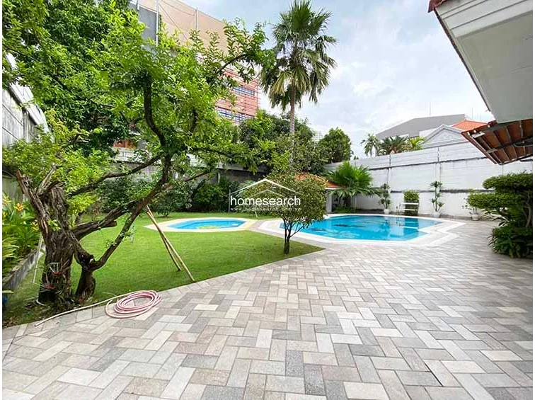 This view highlights the expansive backyard, showcasing a manicured green lawn, a secondary circular wading pool, and a paved stone patio that connects the main residence to the garden. The property is well-maintained with mature trees, shrubs, and a covered gazebo structure, creating a serene and private outdoor retreat.