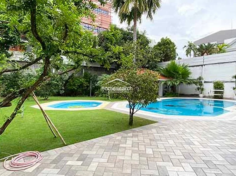 This outdoor space features a well-maintained lawn and a private swimming pool with a secondary smaller pool or spa area. The area is enclosed by a high perimeter wall for privacy and includes mature trees and paved walkways, suggesting a quiet and relaxed environment.