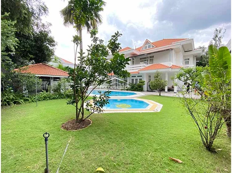 This exterior shot captures the rear elevation of the two-story villa, which features a traditional orange-tiled roof and a white facade. The property includes a private swimming pool, a landscaped garden with a sprinkler system, and a small shaded pavilion, all reflecting a high level of maintenance and architectural consistency.