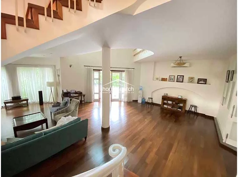 This expansive open-plan living area features high ceilings and a grand staircase, creating a dramatic sense of scale and architectural interest. The room is anchored by rich hardwood flooring and flows seamlessly toward large windows and glass doors that provide abundant natural light and exterior access.