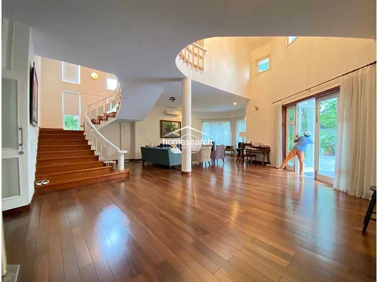 This expansive living area features a grand, double-height ceiling and a striking curved staircase with a white balustrade. The space is finished with polished hardwood flooring and large glass doors that provide abundant natural light and direct access to the outdoor area.