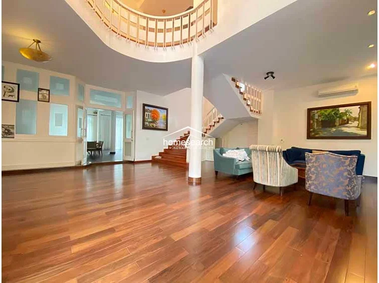 This open-plan living room showcases a dramatic architectural design with a mezzanine-level balcony overlooking the main space. The room is anchored by polished hardwood floors and features a comfortable seating arrangement, with large windows and glass doors ensuring a bright, airy flow.