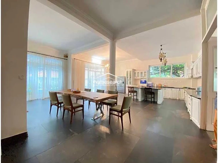 This spacious open-concept area integrates a dining room and kitchen, featuring large dark floor tiles and a natural wood dining table. The kitchen is equipped with white cabinetry and a central island, while expansive windows and sheer curtains allow for excellent natural light throughout the room.