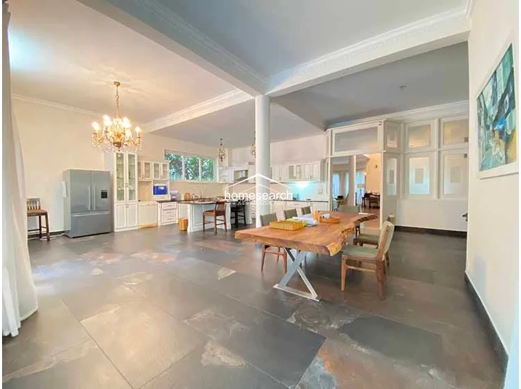 This expansive open-plan living and dining area is defined by high ceilings and a sophisticated, neutral color palette. The room is anchored by a large, natural-edge wooden dining table and features dark, oversized stone flooring that contrasts elegantly with the white cabinetry.