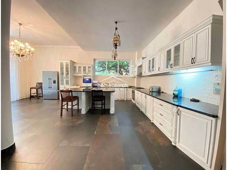 This spacious kitchen features a classic layout with extensive white cabinetry, glass-front display cupboards, and a central island with bar seating. The room is illuminated by an ornate crystal chandelier and recessed lighting, creating a bright and functional workspace with ample storage.
