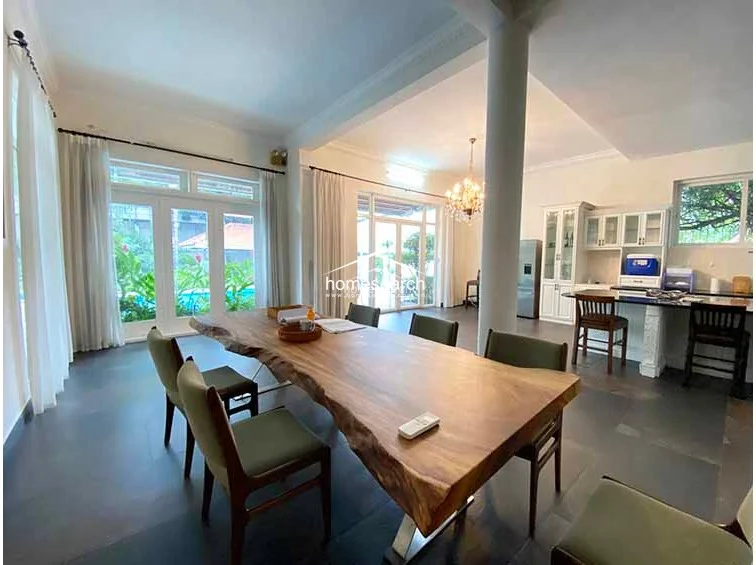 This open-concept dining and kitchen area features a striking live-edge wooden table as the centerpiece, complemented by large floor-to-ceiling windows that provide ample natural light and views of the outdoor pool. The space is anchored by a structural column and defined by dark, large-format floor tiles that contrast beautifully with the crisp white walls and elegant chandelier. The kitchen area includes a breakfast bar with high stools and white cabinetry, maintaining a clean and sophisticated aesthetic throughout the main living level.