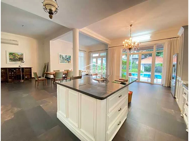 This spacious living and dining area showcases an open floor plan that flows seamlessly toward a private outdoor swimming pool visible through expansive glass doors. The interior is characterized by high ceilings, a grand chandelier, and dark stone flooring, creating a luxurious atmosphere that is both airy and grounded. A large kitchen island with white cabinetry serves as a functional focal point, bridging the gap between the dining space and the rest of the home.