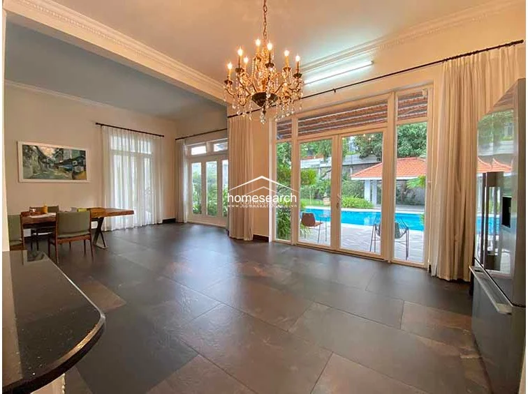 This wide-angle view of the dining and kitchen zone highlights the seamless integration of indoor and outdoor living, with direct access to a sparkling pool area. The room is defined by its dark, durable tile flooring, high ceilings, and a dramatic chandelier that adds a touch of opulence to the functional space. The layout is spacious and uncluttered, emphasizing a bright, airy environment perfect for entertaining.