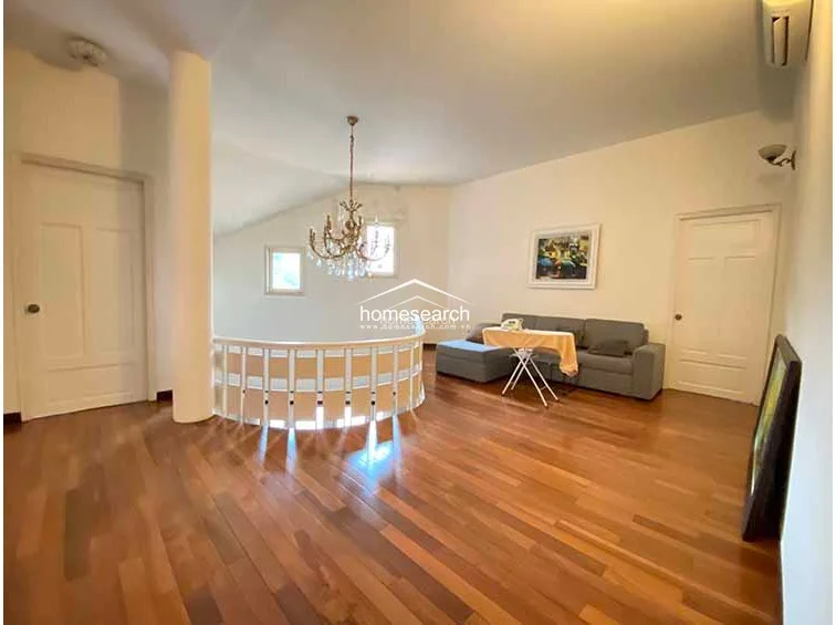 This spacious, open-plan living area features warm hardwood flooring and a unique curved balcony railing overlooking the lower level. The room is illuminated by a grand chandelier and natural light from multiple windows, with a wall-mounted AC unit and a neutral color scheme.