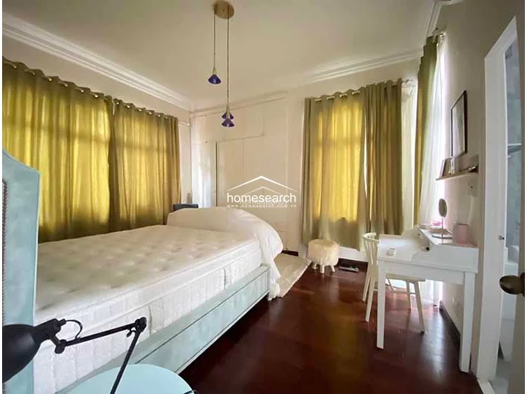 This bedroom is characterized by rich hardwood floors, floor-to-ceiling olive-green curtains, and built-in white wardrobes for ample storage. The space is accented by modern pendant lighting and includes a dedicated white vanity desk, creating a bright and functional sleeping environment.
