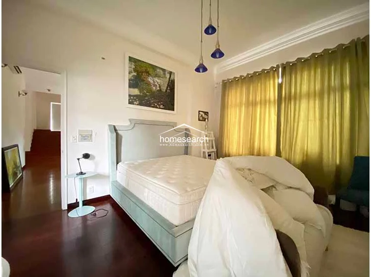 This bedroom features warm hardwood flooring and neutral walls, accented by a decorative blue upholstered bed frame and modern pendant lighting. Large windows dressed in olive-green curtains provide natural light, while an open doorway leads to a staircase, suggesting a multi-level layout.