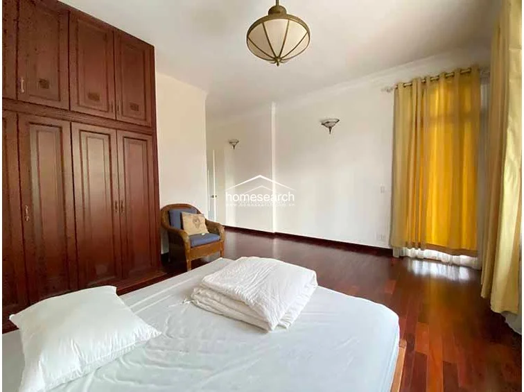 This spacious bedroom maintains a consistent aesthetic with rich hardwood floors, white walls, and a large, dark wood built-in wardrobe for ample storage. A central dome-style light fixture and wall-mounted sconces provide ambient lighting, while the room connects to an adjacent hallway.