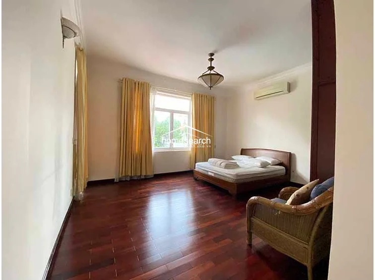 This bedroom offers a clean, minimalist layout with polished hardwood floors and a simple wooden bed frame. The room is well-lit by a large window with yellow curtains and includes a wall-mounted air conditioning unit for climate control.