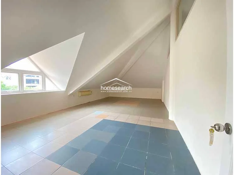 This attic-style room features a unique, sloped ceiling and is finished with light-colored square floor tiles. A large, angular window allows for significant natural light, though the space currently appears empty and serves as a versatile, open-concept area.