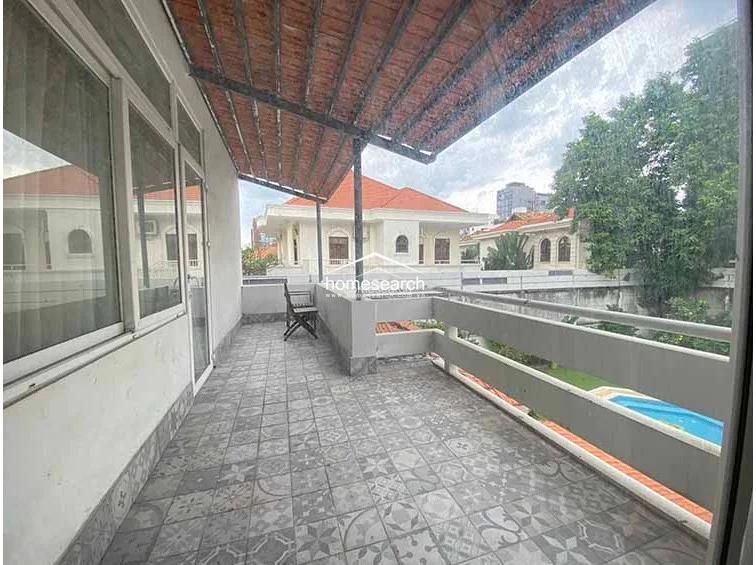 This balcony offers a spacious outdoor retreat with patterned tile flooring and a protective railing overlooking the garden and pool area below. It is partially covered by a rustic, reddish-toned overhead structure, providing a shaded spot for outdoor seating.