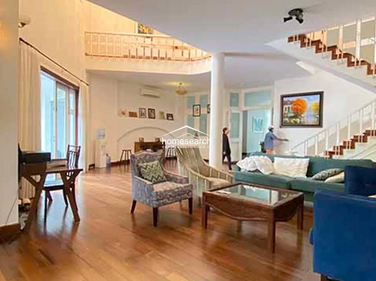 This grand living room is defined by its soaring ceilings, a mezzanine level with a wooden staircase, and warm wood flooring throughout. The space is elegantly furnished with a blue sofa and patterned armchairs, and it features a large wall-mounted painting and a central column that anchors the open-plan layout.
