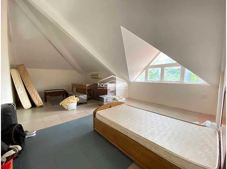 This attic bedroom features a unique slanted ceiling and a large triangular window that brings in natural light. The room is sparsely furnished with a single bed and some storage items, utilizing a neutral color palette and tile flooring.