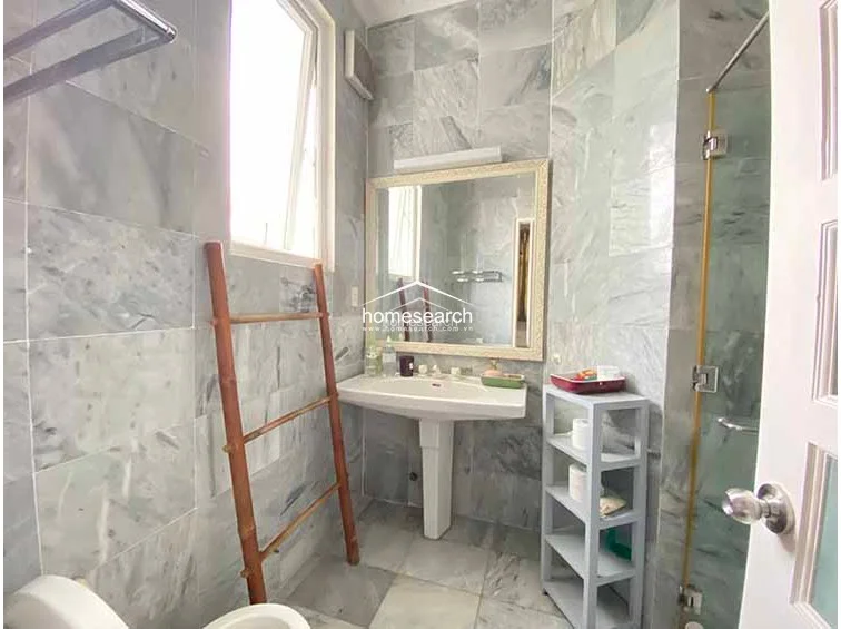 This bathroom is fully finished with grey marble-style wall and floor tiles, giving it a clean and modern aesthetic. It includes a pedestal sink with a framed mirror, a glass-enclosed shower stall, and a small bamboo ladder for towel storage.