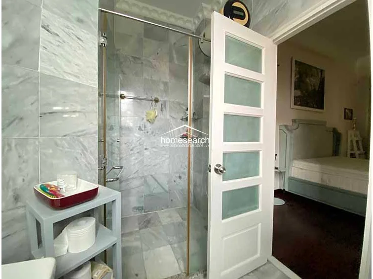 This bathroom features a walk-in shower enclosed by glass with gold-toned fixtures and floor-to-ceiling marble tiling. A white, multi-panel frosted glass door provides privacy while allowing light to filter through, connecting the space directly to the adjacent bedroom.