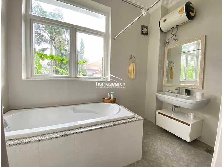 This bright bathroom is centered around a spacious white soaking tub positioned beneath a large window that provides natural light and a view of greenery. The room is finished with neutral-toned walls, marble-style flooring, and a modern wall-mounted vanity, complete with a water heater unit.