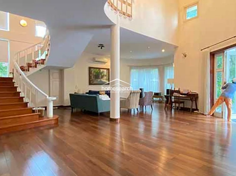 This expansive living area features a dramatic double-height ceiling and a grand curved wooden staircase that serves as a central architectural focal point. The space is finished with polished hardwood flooring and neutral walls, creating an open, airy flow that connects seamlessly to the adjacent dining and kitchen zones.