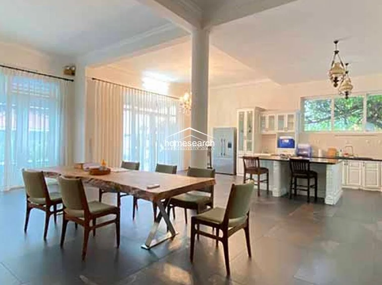 This open-concept dining and kitchen space features a large, rustic live-edge wooden table that anchors the room, set against a backdrop of large windows draped in floor-length curtains. The kitchen is equipped with white cabinetry, a central island with bar seating, and modern appliances, all set upon durable, large-format grey tile flooring.