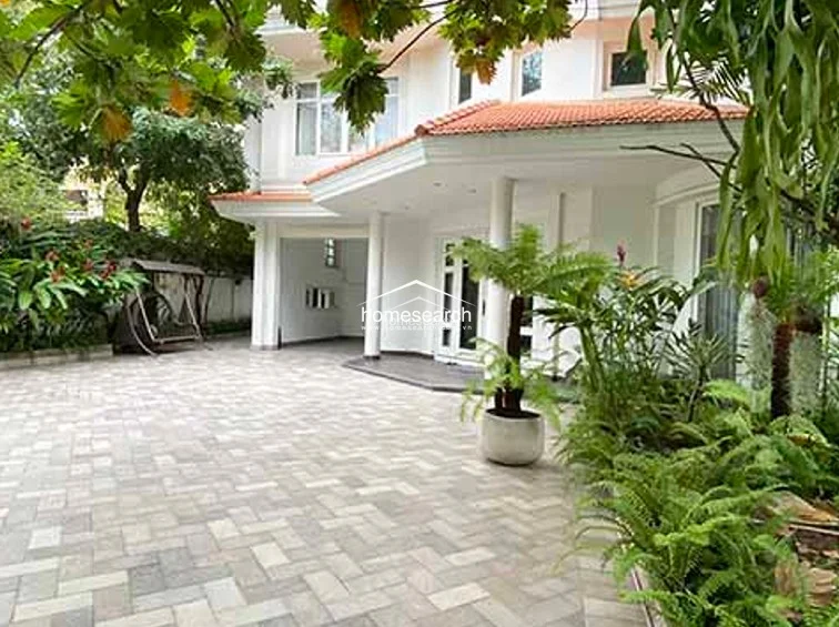 The exterior of the property showcases a well-maintained white facade with classic architectural columns and a paved driveway area. Lush greenery and potted plants frame the entrance, providing a welcoming outdoor space that leads into the home through large, glass-paned doors.