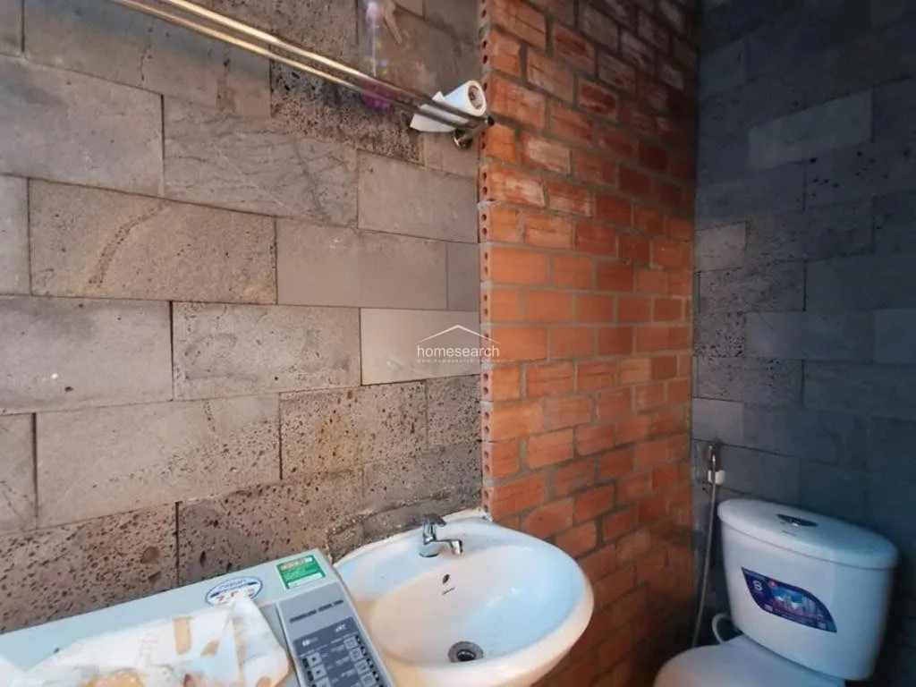 This bathroom features a rustic, industrial aesthetic characterized by a mix of textured grey stone wall cladding and an exposed red brick accent wall. The space is equipped with a standard white pedestal sink, a toilet with a handheld bidet sprayer, and a top-loading washing machine positioned directly beneath the sink area. The overall condition suggests a utilitarian design, though the exposed brick and stone finishes provide a distinct, raw architectural character.