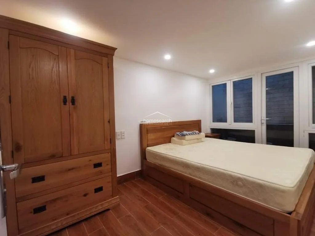 This bedroom features warm wood-look flooring and a matching wooden bed frame and wardrobe, creating a cohesive and cozy aesthetic. The room is well-lit by recessed ceiling lights and includes a large window with white frames that provides natural light and a view of the exterior.