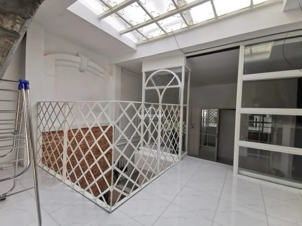This versatile upper-level space acts as a bright atrium or hallway, characterized by a large overhead skylight that floods the area with natural light. It features a decorative white metal lattice railing and a glass-enclosed section, showcasing a clean, modern aesthetic with white tile flooring.