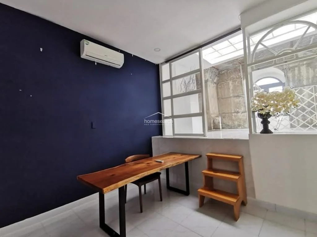 This room serves as a minimalist study or dining area featuring a striking navy blue accent wall and a natural wood slab table. It is equipped with a wall-mounted AC unit and connects to an adjacent space through a large, light-filled glass partition that allows for an airy, open-concept feel.