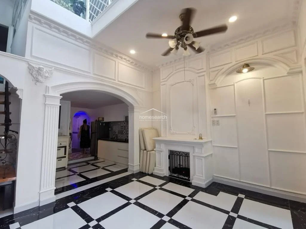This elegant living area is defined by high ceilings, intricate white wall molding, and a dramatic black-and-white tiled floor. The space features a ceiling fan with integrated lighting, a decorative fireplace mantle, and an arched entryway that provides a seamless flow into the kitchen area.