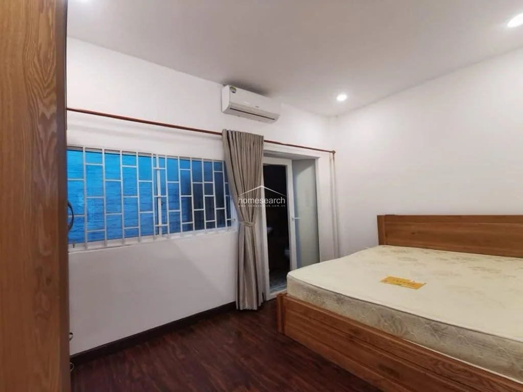 This bedroom maintains a consistent design language with dark wood-look flooring and a sturdy wooden bed frame. It is climate-controlled with a wall-mounted AC unit and features a large window secured with metal bars, allowing for ventilation while maintaining security.