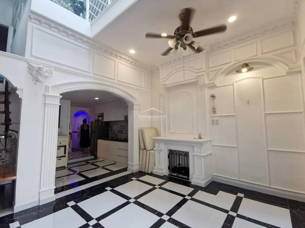 This elegant living area features high ceilings with ornate molding, a central ceiling fan, and a decorative fireplace mantel. The space is finished with high-contrast black and white marble-style flooring and flows directly into the kitchen through a wide, arched entryway.