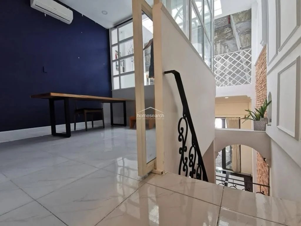 This upper-level landing or study area is defined by a striking navy blue accent wall and bright white marble flooring. It features a modern wooden desk, a wall-mounted AC unit, and a glass-paneled partition that overlooks the lower level, creating an open and airy architectural feel.