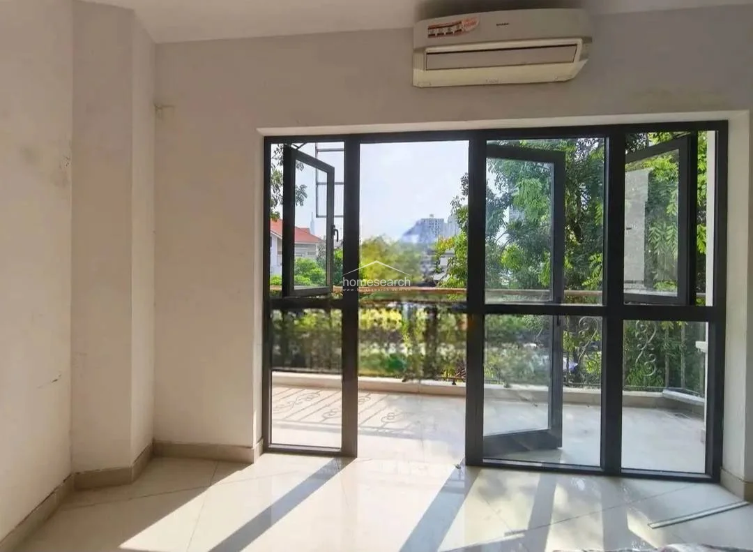 This interior room features a clean, minimalist layout with neutral wall finishes and light-colored tile flooring that enhances the sense of space. A large, dark-framed glass door system provides abundant natural light and seamless access to an outdoor balcony, while a wall-mounted AC unit is included for climate control.