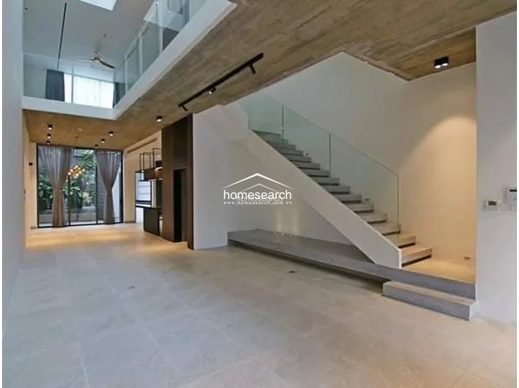 This spacious, open-concept living area features a dramatic double-height ceiling with a raw concrete finish and a sleek glass-railed staircase. The room is finished with large-format light-colored floor tiles and flows directly into the kitchen, illuminated by both natural light from the pool-facing windows and modern track lighting.