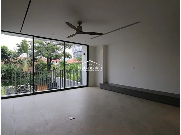 This spacious, minimalist living area features large floor-to-ceiling sliding glass doors that provide abundant natural light and a lush, green view. The room is finished with neutral grey floor tiles, white walls, and a modern ceiling fan, complemented by a low-profile, dark-toned built-in media console.