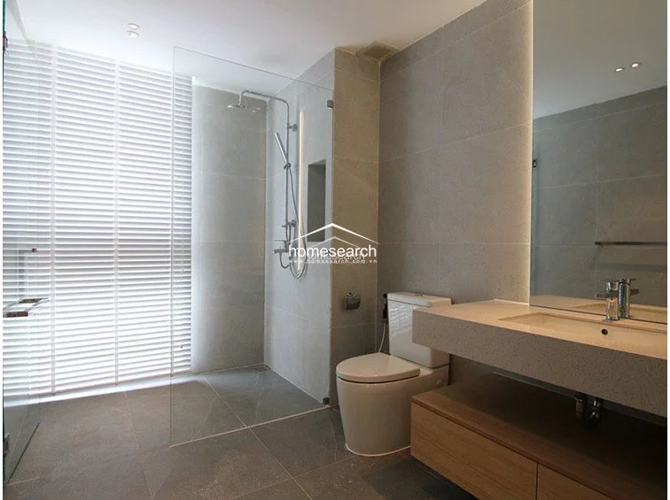 This modern bathroom features a sleek glass-enclosed walk-in shower, a floating vanity with a stone countertop, and a wall-mounted toilet. The room is finished in neutral grey stone tiles and utilizes white Venetian blinds to manage natural light while maintaining privacy.
