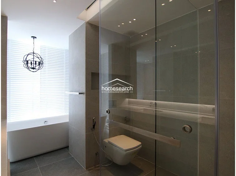 This luxurious bathroom is designed with a contemporary aesthetic, featuring a freestanding soaking tub positioned beneath a decorative modern chandelier. It includes a glass-enclosed shower, a wall-mounted toilet, and high-quality grey stone tiling throughout, creating a spa-like atmosphere.