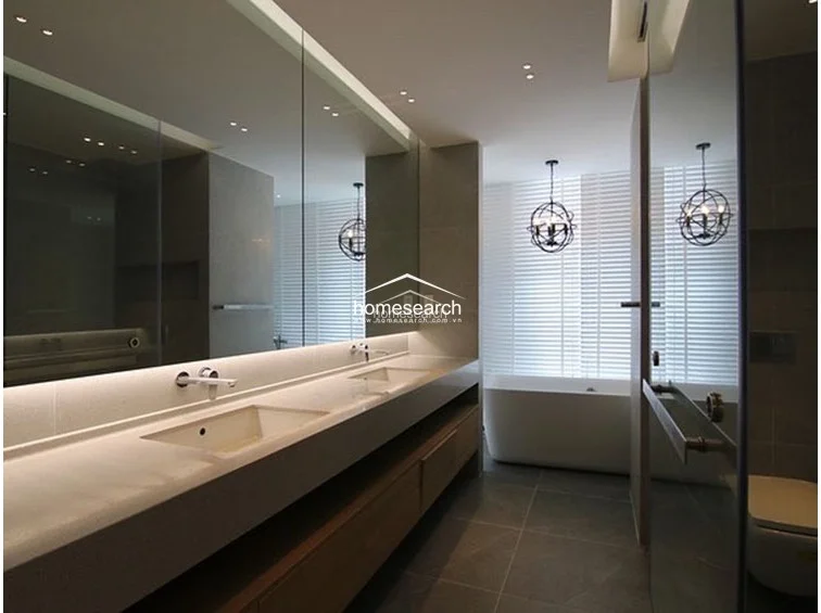 This modern, spa-inspired bathroom features a sleek double vanity with integrated sinks, a large wall-to-wall mirror, and warm under-cabinet LED lighting. The space includes a deep soaking tub positioned by a window with adjustable blinds for natural light, complemented by high-end dark floor tiles and a minimalist, neutral color palette.