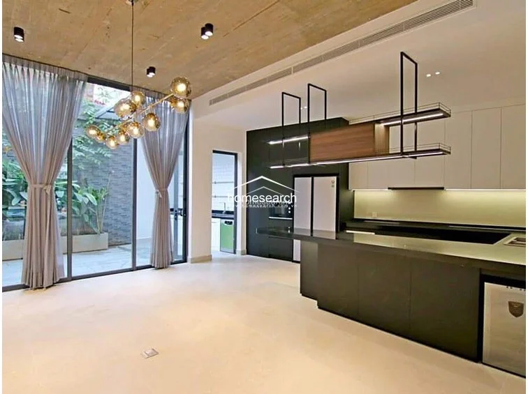 The kitchen is a sophisticated, modern space featuring a large island, dark cabinetry, and a unique suspended metal shelving unit with integrated lighting. It connects fluidly to the living area, where a decorative multi-globe chandelier hangs near the floor-to-ceiling glass doors that look out onto the lush outdoor greenery.