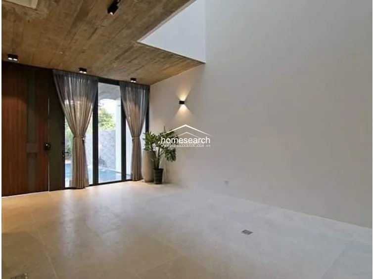 This minimalist living room area emphasizes vertical space with its double-height void and clean, white walls accented by a warm, modern wall sconce. The room is anchored by large-format floor tiles and features a dark wood door, with a large potted plant adding a touch of nature near the expansive glass doors leading to the pool.