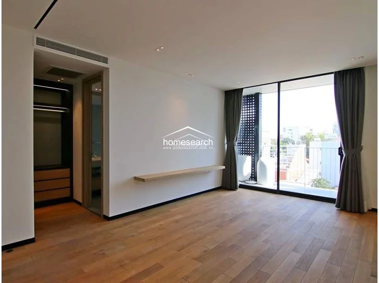 This bedroom features warm wood flooring and a minimalist aesthetic, highlighted by a floating wooden shelf and a built-in open-concept wardrobe system. Large sliding glass doors provide access to a private balcony with a metal railing, offering an elevated view of the surrounding neighborhood and ensuring the room is filled with natural light.